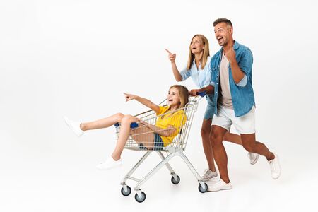 Image of happy family man and woman having fun while giving ride to their daughter in shopping cart isolated over white backgroundの写真素材