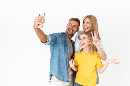 Photo of happy woman and man with little girl smiling while taking selfie on cellphone isolated over...