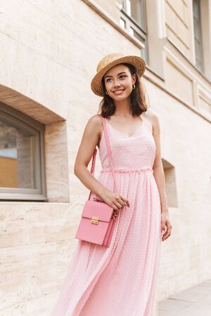 Photo of beautiful young woman wearing summer dress and straw hat smiling while walking on city streetの写真素材