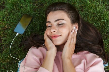 Top view close up of a lovely young brunette girl laying on grass outdoors, listening to music with...