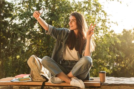 Image of adorable brunette woman smiling and taking selfie photo on smartphone while sitting at bench in green park on summer dayの写真素材
