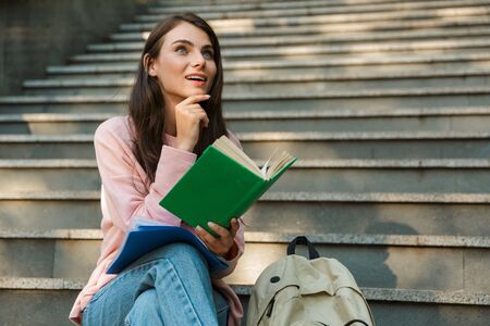 Attractive young smiling girl student reading a book while sitting on staircaseの写真素材