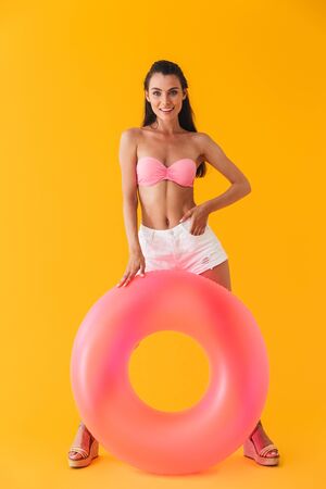 Full length of an attractive cheerful young girl wearing bikini standing with inflatable ring isolated over yellow background, posingの写真素材