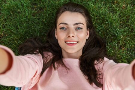 Top view close up of a lovely smiling young brunette girl laying on grass outdoors, showing tongue,...