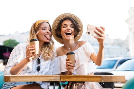 Photo of a cutie happy smiling young optimistic girls friends sitting outdoors in cafe drinking coffee take selfie by mobile phone showing tongue.の写真素材