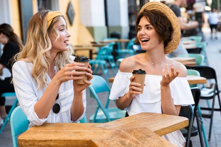 Image of a cheery young girls friends sitting outdoors in cafe drinking coffee talking with each other.の写真素材