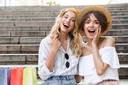 Image of a surprised emotional happy young girls friends sitting outdoors on steps hugging each other near shopping bags.の写真素材