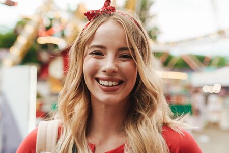 Image of pretty blonde woman wearing girlish clothes smiling and standing in front of colorful carousel at amusement parkの写真素材