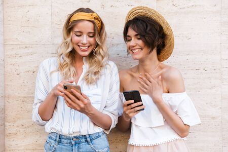 Image of a cheery pleased young women friends outdoors using mobile phones chatting.の写真素材