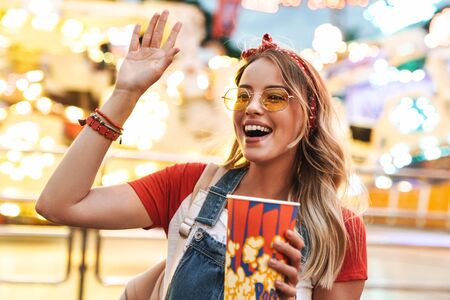 Image of cheerful blonde woman wearing girlish clothes waving and holding popcorn while walking in amusement parkの写真素材