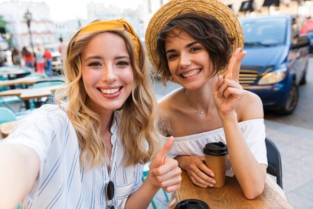 Photo of a smiling positive young women friends sitting outdoors in cafe drinking coffee take a selfie by camera showing peace and thumbs up.の写真素材