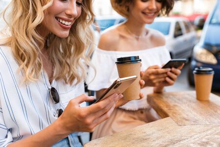 Cropped photo of a cheerful young pleased optimistic girls friends sitting outdoors in cafe drinking coffee using mobile phones.の写真素材