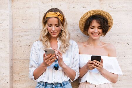 Image of a smiling pleased happy positive young women friends outdoors using mobile phones chatting.の写真素材