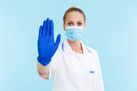 Beautiful angry blonde woman doctor wearing uniform and mask standing isolated over blue background, showing stop gestureの写真素材