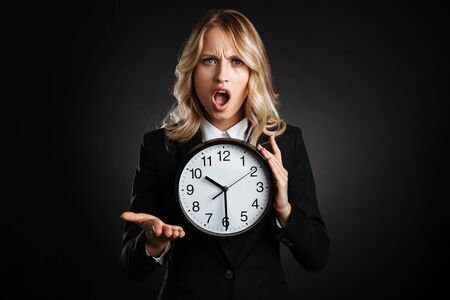 Portrait of a beautiful shocked blonde haired business woman dressed in formal clothes standing isolated over black background, showing alarm clockの写真素材
