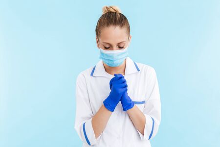Beautiful blonde woman doctor wearing uniform and mask standing isolated over blue background,...