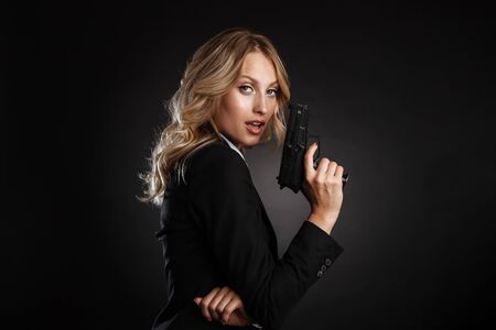 Portrait of a beautiful blonde haired business woman dressed in formal clothes standing isolated over black background, shooting with a gunの写真素材