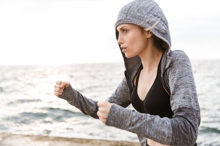 Image of serious blonde woman wearing hoodie boxing while working out on pier near seaside in morningの写真素材
