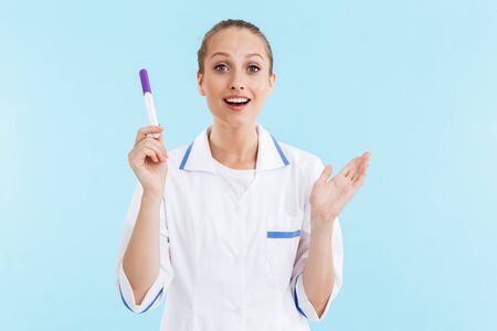 Beautiful excited blonde woman doctor wearing uniform standing isolated over blue background, showing pregnancy testの写真素材