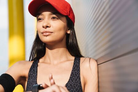 Attractive young fitness girl leaning on a wall while standing outdoors, checking smart watchの写真素材