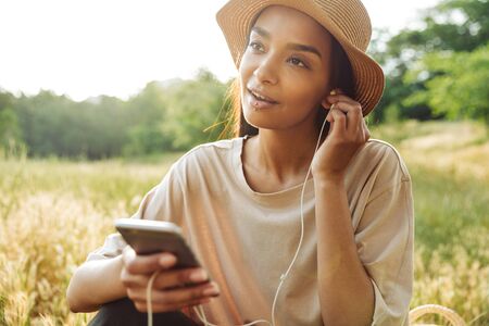 Portrait of pretty woman wearing lip piercing and straw hat listening to music on smartphone while sitting on grass in green parkの写真素材