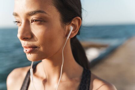 Portrait of pleased strong sports woman outdoors at the beach at morning posing listening music with earphones.の写真素材