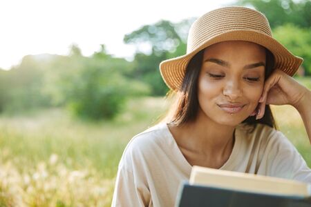 Portrait of satisfied woman wearing lip piercing and straw hat reading book while sitting on grass in green parkの写真素材
