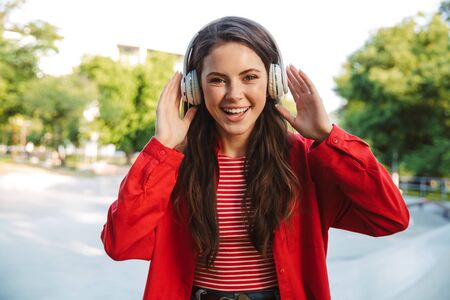 Image of laughing girl student wearing red jacket listening music with headphones on sports ground