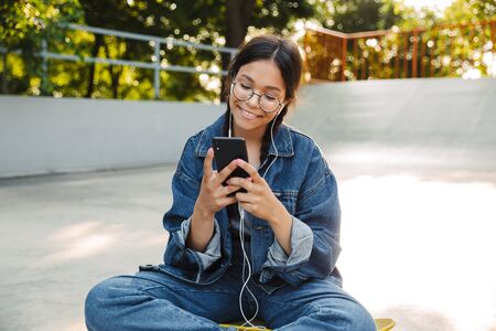 Image of pretty girl dressed in denim wear using smartphone and earphones while sitting on skateboard in skate parkの写真素材