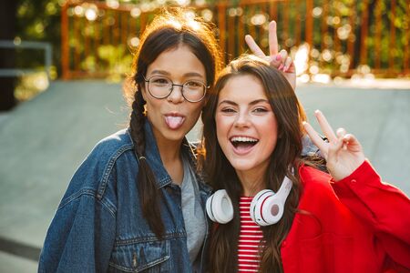 Image of two amusing girls dressed in denim wear laughing and showing peace sign while walking in parkの写真素材