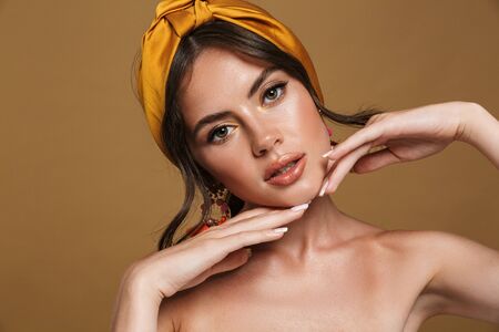 Close up beauty portrait of an attractive young topless woman wearing headband standing isolated over brown background, posing, touching her faceの写真素材