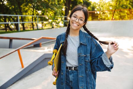 Image of caucasian girl dressed in denim wear smiling and rejoicing while holding skateboard in skate parkの写真素材