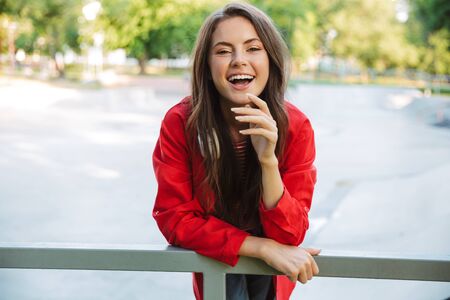 Image closeup of pleased girl student wearing red jacket laughing and bending on railing on sports groundの写真素材