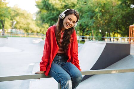 Image of joyful girl student wearing red jacket listening music with headphones while sitting on railing at sports groundの写真素材