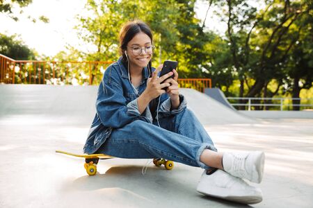 Image of beautiful girl dressed in denim wear using smartphone and earphones while sitting on skateboard in skate parkの写真素材