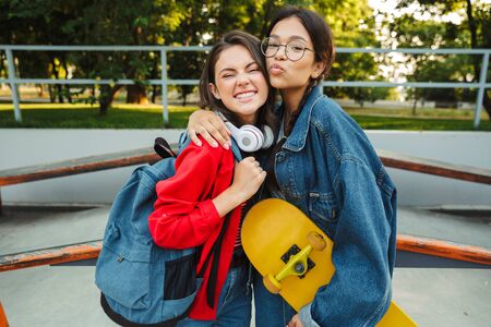 Image of two cute girls dressed in denim wear smiling and hugging together while holding skateboard in skate parkの写真素材