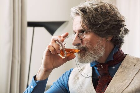 Image of handsome adult professor man wearing stylish suit and eyeglasses drinking alcohol from glass while sitting on armchair in hotel apartmentの写真素材