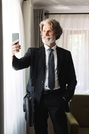 Bearded adult businessman wearing black suit taking selfie on smartphone while standing near window in hotel apartmentの写真素材