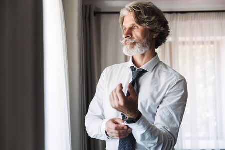 Image of a serious concentrated bearded mature handsome grey-haired businessman posing indoors at home near window dressed in formal clothes correct his cuff links.の写真素材