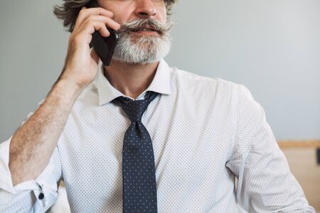 Image closeup of bearded adult man wearing suit and tie talking on cellphone while sitting on...