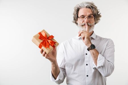 Portrait of a handsome mature businessman dressed in formal wear standing isolated over white background, holding present box, showing silence gestureの写真素材