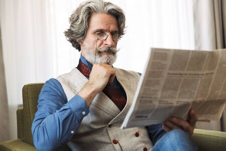 Portrait of smart adult businessman wearing stylish suit reading newspaper while sitting on armchair...