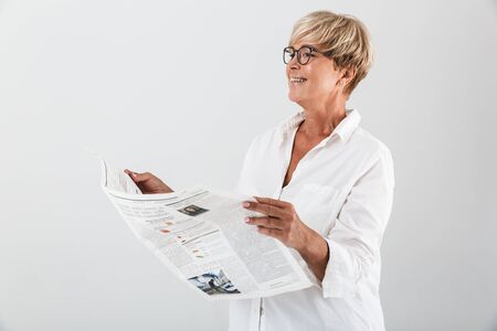 Portrait of happy adult woman wearing eyeglasses smiling and reading newspaper isolated over white background in studioの写真素材