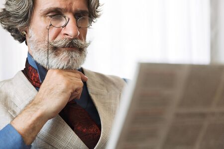 Portrait of concentrated adult businessman wearing stylish suit reading newspaper while sitting on...