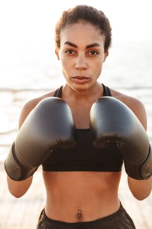 Portrait of fitness pretty woman wearing sportive clothes training in boxing gloves by seaside in morningの写真素材