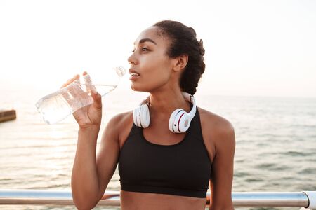 Image of beautiful woman in sportive clothes with headphones bending over railings and drinking water by seaside in morningの写真素材