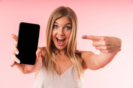 Image of amazed young woman wearing dress holding smartphone and pointing at black screen isolated over pinkの写真素材