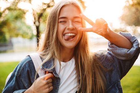 Image of a happy positive young teenage girl walking outside in nature green park showing tongue and peace gesture.の写真素材