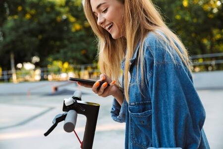 Image of a happy positive smiling cheerful teenage girl in park walking on scooter using mobile...