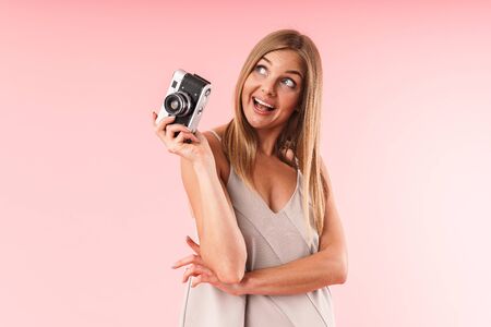 Image of attractive blond woman wearing dress smiling while holding retro camera isolated over pink  in studioの写真素材